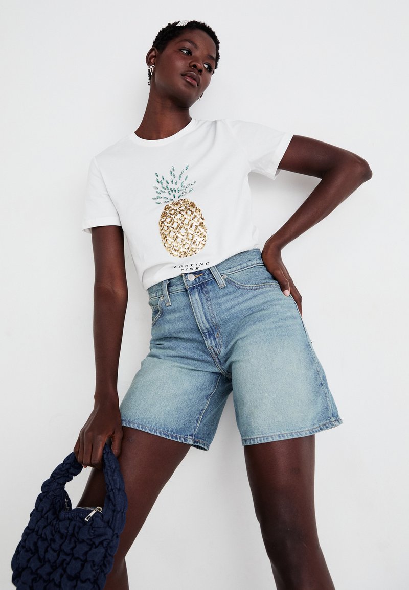 T-shirt blanche avec un design d'ananas doré et bleu, accompagné d'un short en denim bleu clair. Le modèle tient un sac à main plissé marine.