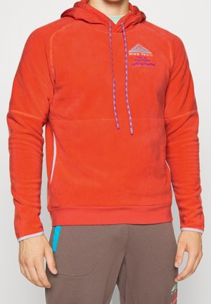 Mand iført en orange hættetrøje i fleece med lilla snore og brune bukser med turkis lommebånd, stående mod en ensfarvet baggrund.