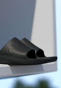 Black slide sandals with open toes, smooth matte finish, curved design, featuring a logo accent on the side and a textured sole for grip.