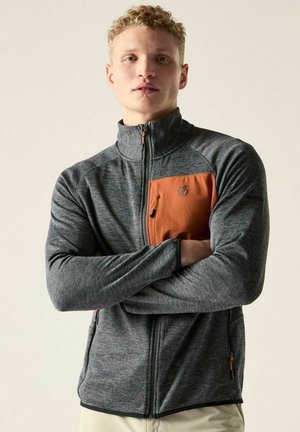 Junger Mann mit lockigem blondem Haar, der eine graue und orange Zip-up-Jacke trägt und die Arme vor einem einfarbigen Hintergrund verschränkt hat.