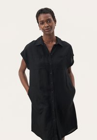 Abito camicia in lino nero con maniche corte, chiusura a bottoni, due tasche sul petto e tasche laterali. Vestibilità comoda e lunghezza al ginocchio.