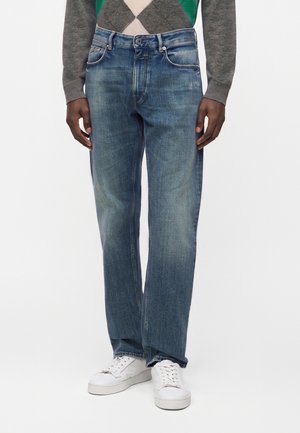 Homme portant un jean droit bleu délavé, des baskets blanches et un pull gris avec un motif losange vert et beige.