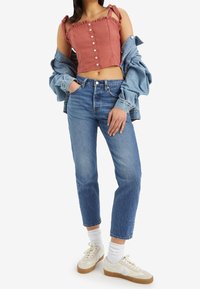 Top de tela rosa recortado con bordes rizados y detalles de botones, combinado con jeans de tiro alto azul y zapatillas blancas con suela marrón.