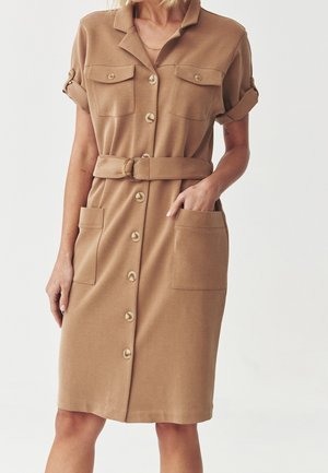 Femme portant une robe-chemise couleur beige avec manches retroussées, deux poches poitrine, deux grandes poches avant et une ceinture assortie à la taille.