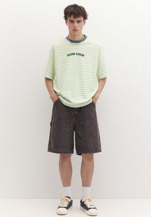 Jeune homme les mains dans les poches porte une chemise "SLOW CREW" rayée verte, un short sombre jusqu'aux genoux, des chaussettes blanches et des baskets noires devant un mur blanc.