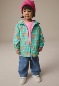Veste légère à carreaux verts avec des motifs arc-en-ciel multicolores, associée à une chemise rose, un jean large en denim et des baskets blanches.