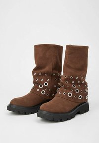 Bottes marron mi-mollet avec des semelles noires épaisses, décorées de plusieurs tailles d'œillets en métal argenté autour de la cheville.