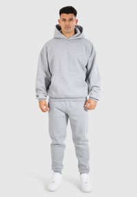 Grauer Hoodie mit Kängurutasche und Kapuze mit Kordelzug, kombiniert mit passenden grauen Jogginghosen. Das Material wirkt weich und strukturiert. Weiße Sneakers.