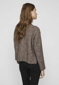 Brūns adīts džemperis ar brīvu piegriezumu, īsu apakšmalu un teksturētu audumu. Garas piedurknes ar ribinātiem uzmaviem. Valkāts kopā ar melnajām biksēm.