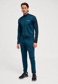 Teal sportieve trainingspak van lichtgewicht, gladde stof met een halve ritskraag. Zwarte accenten op de mouwen. Gestyled met zwarte sportschoenen.
