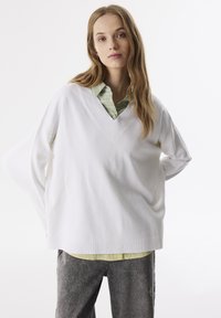 Bílé oversized svetr s V výstřihem a ribbovaným lemem, zkombinovaný se zelenou košilí s límečkem. Měkká, hladká textura; casual, uvolněný střih.