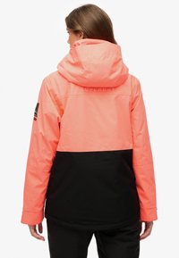 Jasnoróżowa i czarna wodoodporna kurtka z kapturem, z teksturowanym wykończeniem, logo z przodu oraz regulowanymi mankietami dla pewnego dopasowania.