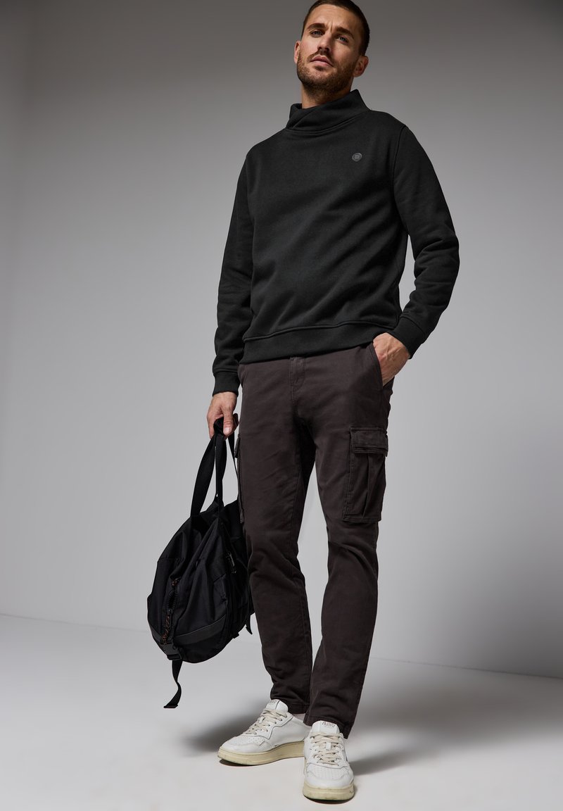 Svart sweatshirt med hög krage, mörka cargobyxor och vita sneakers. Mannen håller en svart väska; släta texturer och enkel design.