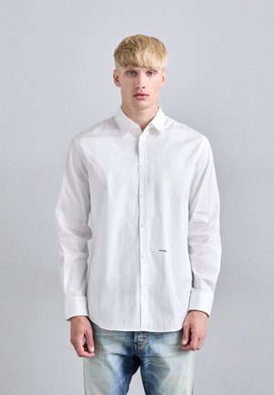 Chemise blanche à col classique, à manches longues, avec un petit logo sur le bas à l'avant, associée à un jean en denim bleu clair.
