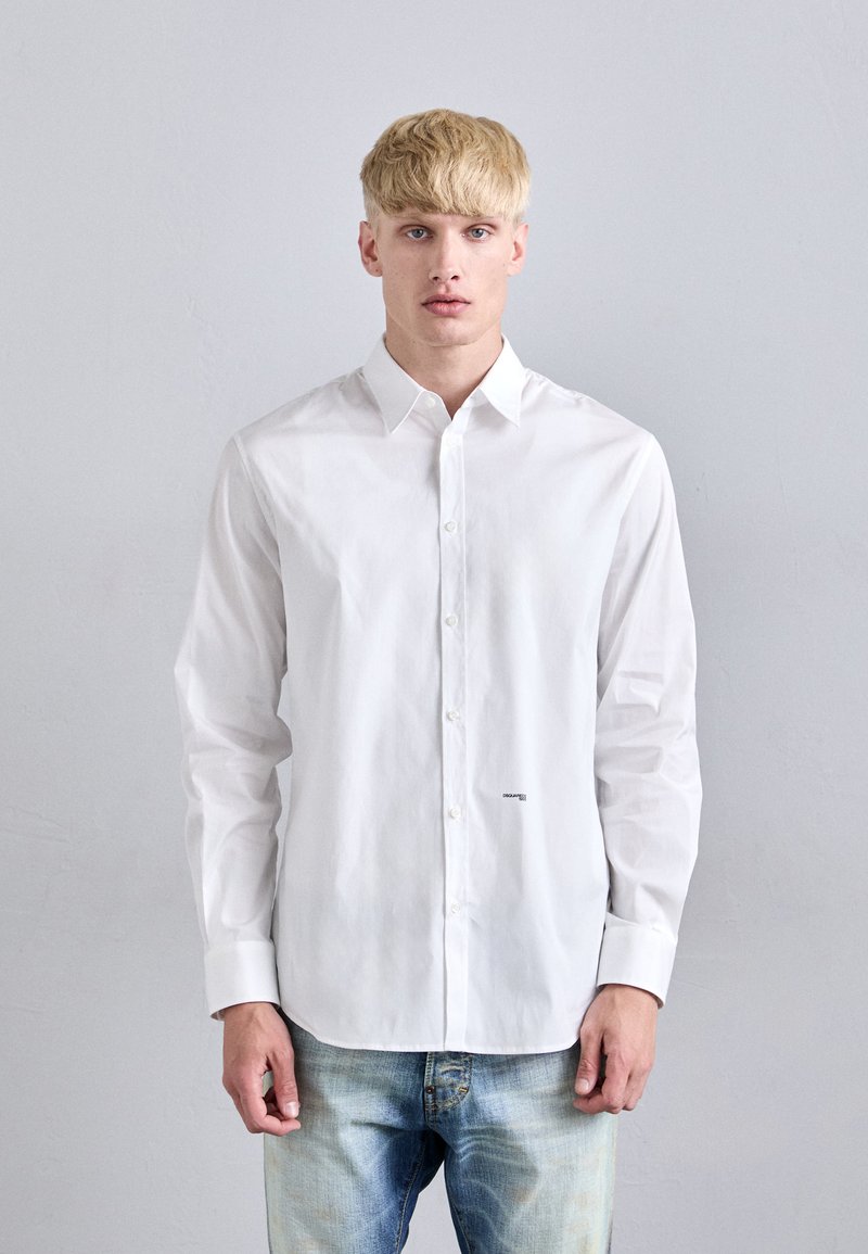 Chemise blanche à col classique, à manches longues, avec un petit logo sur le bas à l'avant, associée à un jean en denim bleu clair.