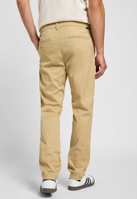 Chinos beige de mezcla de algodón con una textura suave. Presenta dos bolsillos traseros con cierre de cremallera, un diseño ajustado y puños en los tobillos.