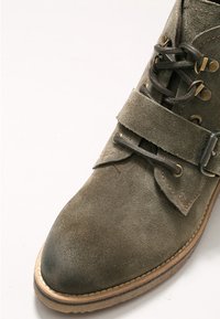 Bottine en daim marron à bout rond, lacets noirs, œillets métalliques, large sangle avec boucle et semelle marron clair.