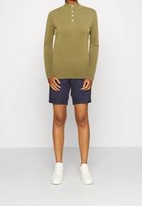 Pull a manica lunga a coste verde oliva con colletto alto e patta con cinque bottoni, abbinato a pantaloni corti blu navy e sneaker bianche.