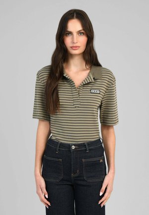 Jeune femme aux longs cheveux bruns portant un polo rayé vert et noir et un jean taille haute bleu foncé sur fond gris.