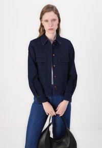 Giacca di jeans scura con chiusura a bottoni, due tasche sul petto e design cropped. Il modello tiene una borsa nera, abbinata a jeans blu.