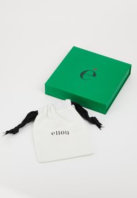 Grøn boks med en glat overflade og et sort logo, ved siden af en beige snørepose med sorte snore og ordet "éliou" trykt på den.