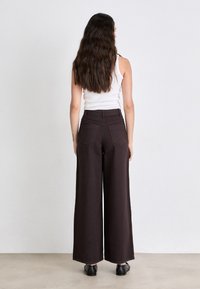 Pantaloni marroni a gamba larga con vita alta, tasche posteriori e una texture liscia, abbinati a un semplice top bianco e scarpe nere.