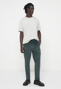 Grijze katoenen t-shirt gecombineerd met gestructureerde, donkergroene broek. Model staat casual met zwarte sneakers aan, handen in de zakken, neutrale achtergrond.