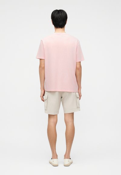 Άνδρας που στέκεται με την πλάτη, φοράει ανοιχτό ροζ μπλουζάκι, μπεζ σορτς cargo και λευκά παπούτσια slip-on σε απλό φόντο.