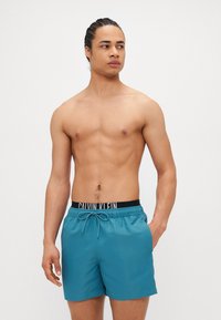 Τουρκοζαρί κολυμβητικά σορτσάκια με ελαστική μαύρη μέση που φέρει το λογότυπο "CALVIN KLEIN", κλείσιμο με κορδόνι και πλευρικές τσέπες. Απαλή υφή.