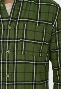 Zielona flanelowa koszula w kratę z zapięciem na guziki, z kontrastującymi czarnymi i białymi liniami oraz kieszenią na piersi po lewej stronie.
