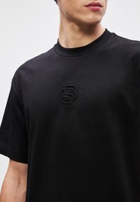 Μαύρη βαμβακερή μπλούζα με κανονική λαιμόκοψη και κοντά μανίκια. Διαθέτει εμποτισμένο λογότυπο στην τσέπη, λεία υφή και άνετη εφαρμογή.