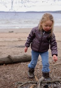 Kind in een paarse bloemenjas en blauwe spijkerbroek verzamelt kleine voorwerpen bij de waterkant, met kale boomtakken en een waterachtergrond.