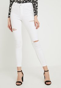 Vaqueros pitillo blancos desgastados con bordes deshilachados, con cintura alta y diseño de cinco bolsillos, combinados con sandalias negras de tacón alto con tiras.