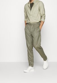 Vaaleanvihreä napitettava paita tummanvihreän T-paidan päällä, yhdistettynä vaaleanharmaisiin joustaviin housuihin ja valkoisiin lenkkareihin neutraalilla taustalla.