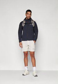 Marineblauwe windbreaker met rits, lichte korte broek en een lichtgekleurde rugzak. Opvallende kenmerken zijn logo-accenten en sportief schoeisel.