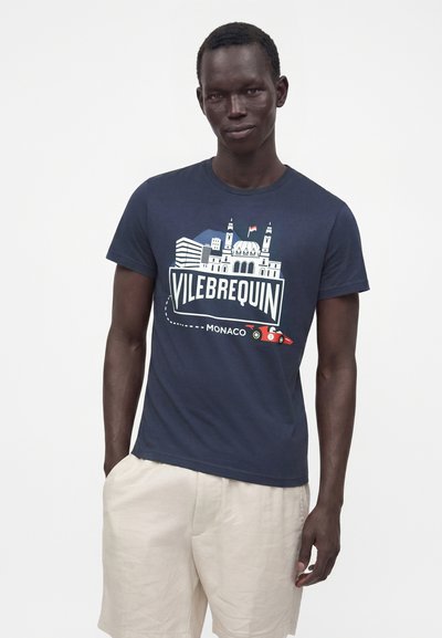 Νέος άνδρας που φοράει μπλε σκούρο T-shirt Vilebrequin Monaco με γραφικά και μπεζ σορτς, στέκεται μπροστά σε λευκό φόντο.