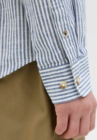 Camicia a righe con linee blu e bianche, polsino aderente con due bottoni beige, abbinata a tessuto khaki visibile sotto la camicia.