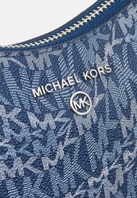 Sininen kuvioitu käsi- tai olkalaukku, joka on valmistettu tekstuuripinnasta. Laukussa on hopeavärinen vetoketju ja metallinen "Michael Kors" -logo, jonka keskellä on pyöreä MK-embleemi.