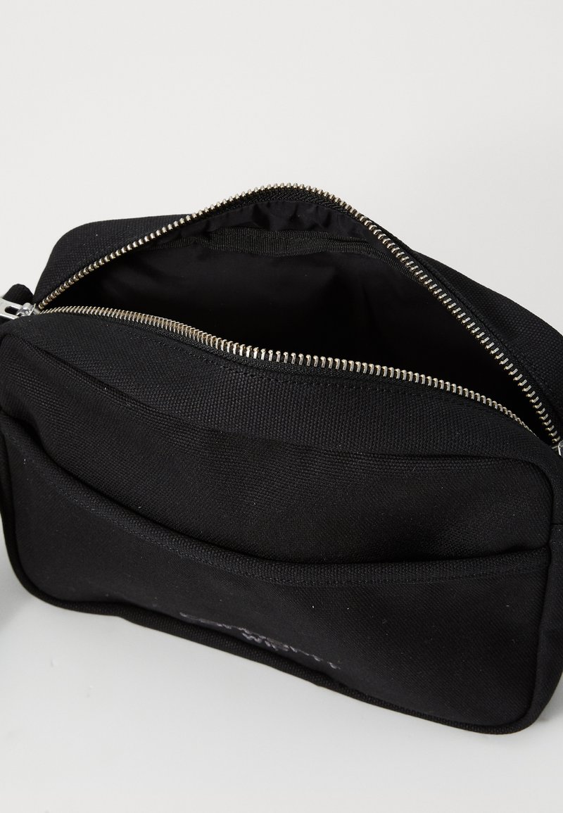 Bolsa de tela negra con una superficie texturizada, parte superior sin cremallera y una cremallera plateada. Presenta un bolsillo frontal y un diseño minimalista.