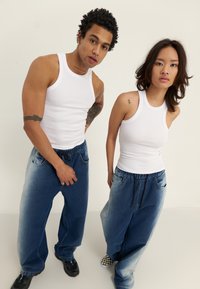 Două persoane poartă tricouri fără mâneci, albe, cu dungi și blugi largi, decolorati, cu snur. Hainele au o croială lejeră și sunt stil casual.