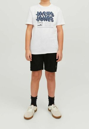Niño de pie con camiseta blanca "JACK & JONES", pantalones cortos negros, calcetas negras y zapatillas blancas sobre un fondo claro liso.