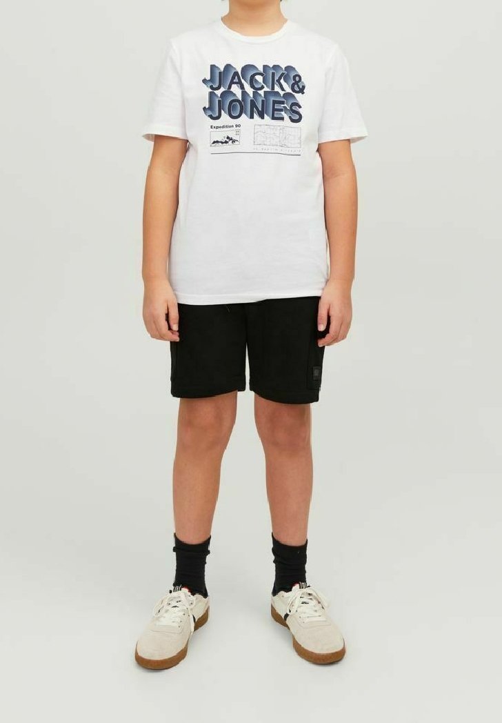 Enfant debout portant un t-shirt blanc "JACK & JONES", un short noir, des chaussettes noires et des baskets blanches sur un fond clair uni.