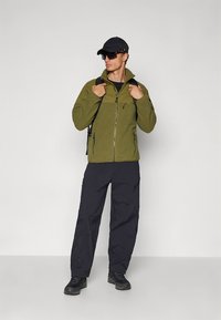 Kurtka polarowa w oliwkowo-zielonym kolorze z zamkiem błyskawicznym z przodu, czarne kieszenie na zamek, zestawiona z czarnymi spodniami i solidnymi butami, noszona z czarną czapką i okularami przeciwsłonecznymi.