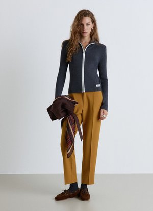 Donna con un maglione scuro con zip, pantaloni color senape e scarpe marroni, che tiene in mano sinistra una giacca piegata con una sciarpa a righe.