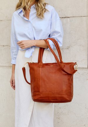 Žena oblečená v svetlomodrej košeli a bielej sukni drží veľkú hnedú koženú tašku s dvoma rukoväťami a ramenným popruhom.