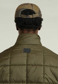 Olivgrön vadderad jacka med hög krage och fyrkantigt quiltmönster. Har en svart logotyp-patch på ryggen. Kamouflagekeps ingår.