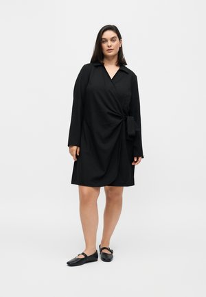 Femme portant une robe portefeuille noire à manches longues avec un nœud sur le côté et des chaussures plates noires, debout devant un fond clair uni.