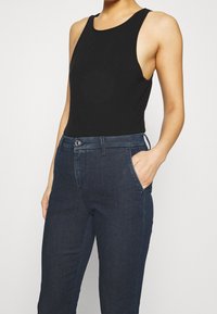 Bodysuit negro sin mangas con escote redondeado, combinado con unos vaqueros de mezclilla azul oscuro que tienen un bolsillo frontal y cierre con botón de metal.