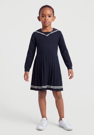 Enfant debout portant une robe plissée bleu marine à manches longues avec des rayures blanches et des baskets blanches sur un fond uni.