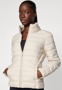 Licht beige gewatteerde jas met een ritssluiting, hoge kraag en zijzakken. Bevat horizontale gestikte lijnen en een gladde textuur.